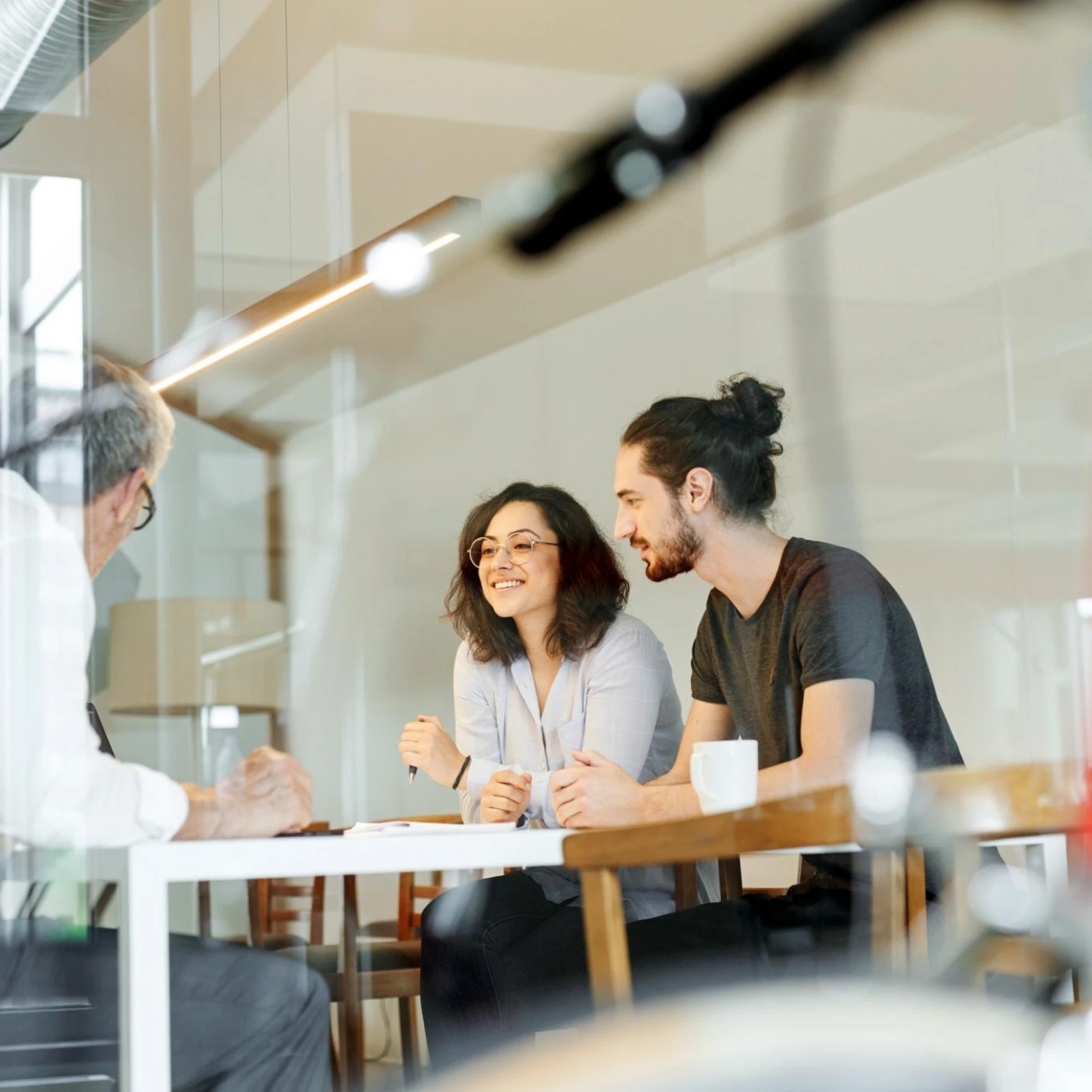 A relaxed business meeting with a man and a woman seated at a table, smiling and engaging in conversation with an older man. The setting is a modern, glass-walled office, with a red bicycle partially visible in the foreground.