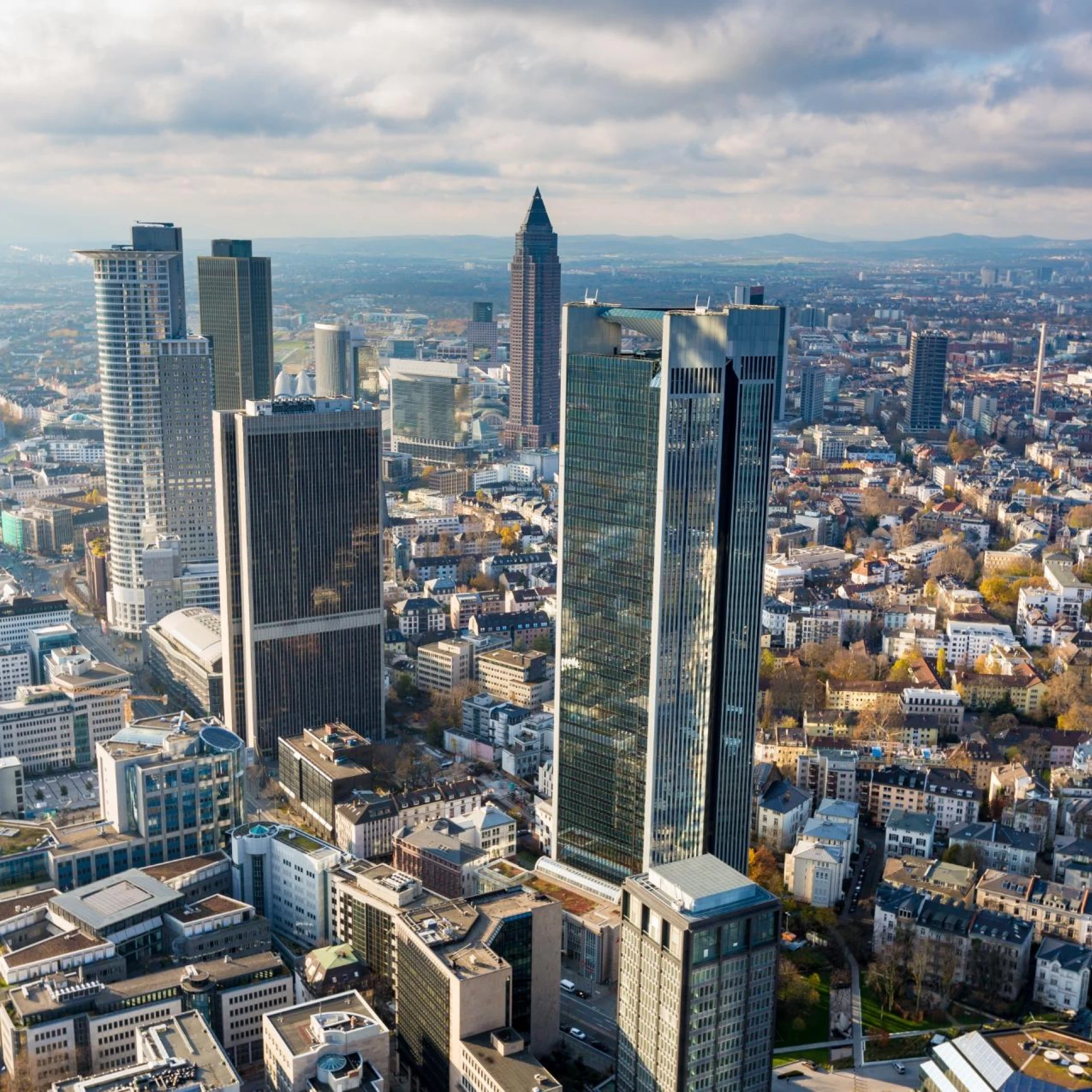 An aerial view of Frankfurt, Germany, highlighting its modern skyscrapers, including the Deutsche Bank towers and Commerzbank Tower, amidst a sprawling urban cityscape.