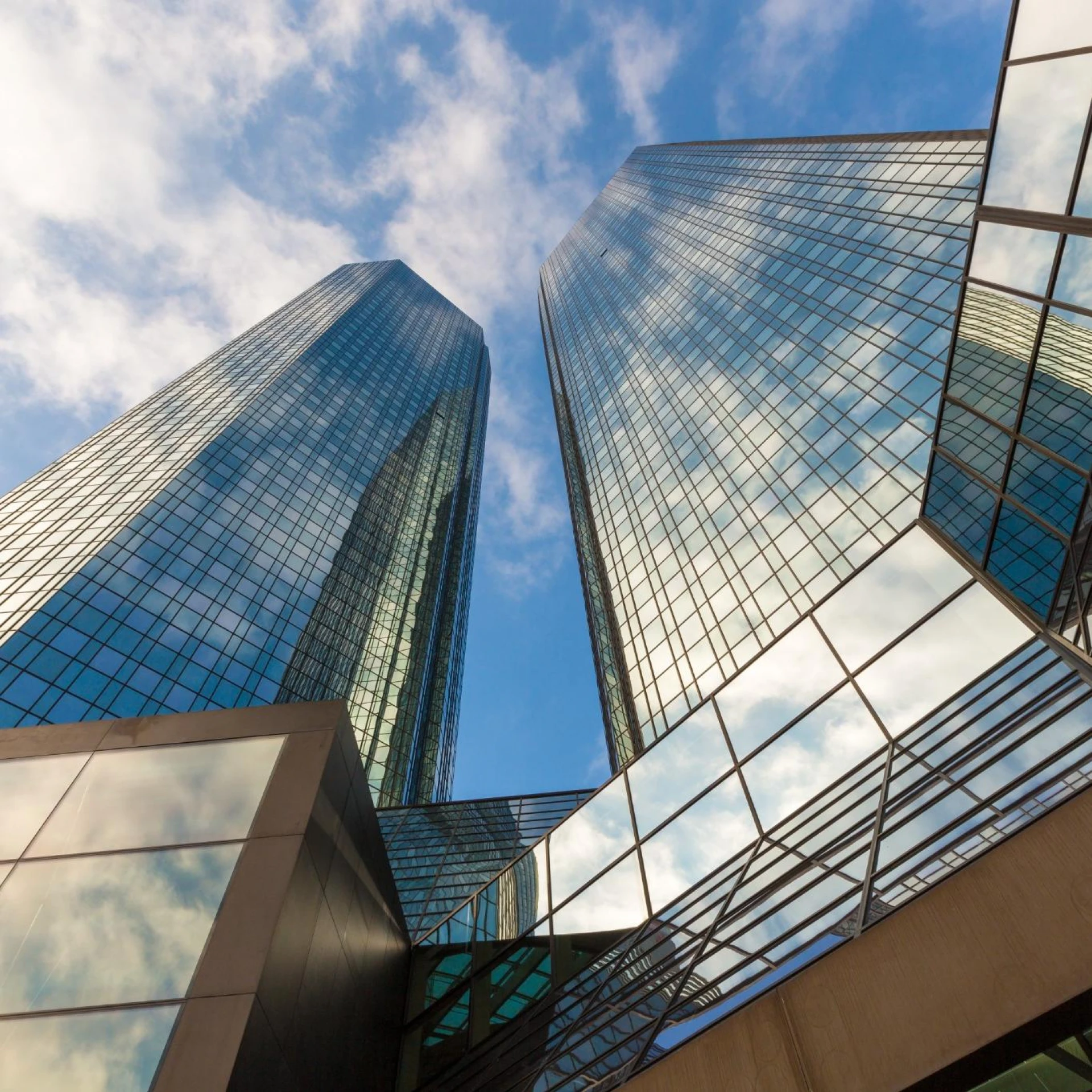 A low-angle view of tall, modern glass skyscrapers reflecting the blue sky and clouds. The geometric architecture emphasizes the sleek, urban design and professional corporate environment.