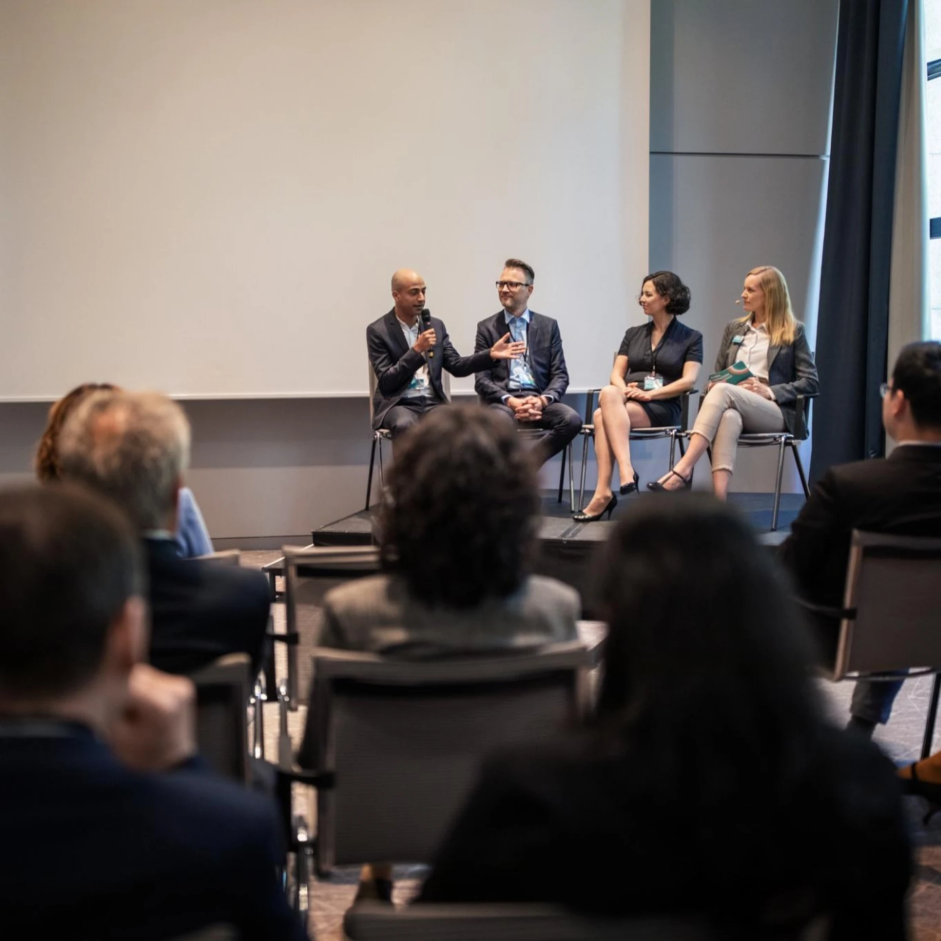 A panel of four speakers seated on stage, engaging with an audience in a conference room. One speaker is holding a microphone while others listen attentively.