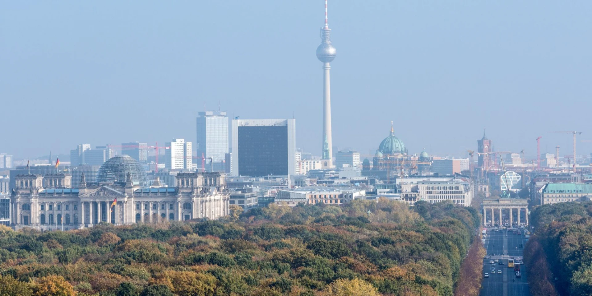 An aerial view of Berlin, Germany, showcasing the iconic landmarks such as the Reichstag building, Berlin TV Tower, and the Brandenburg Gate, surrounded by lush greenery and a clear sky.