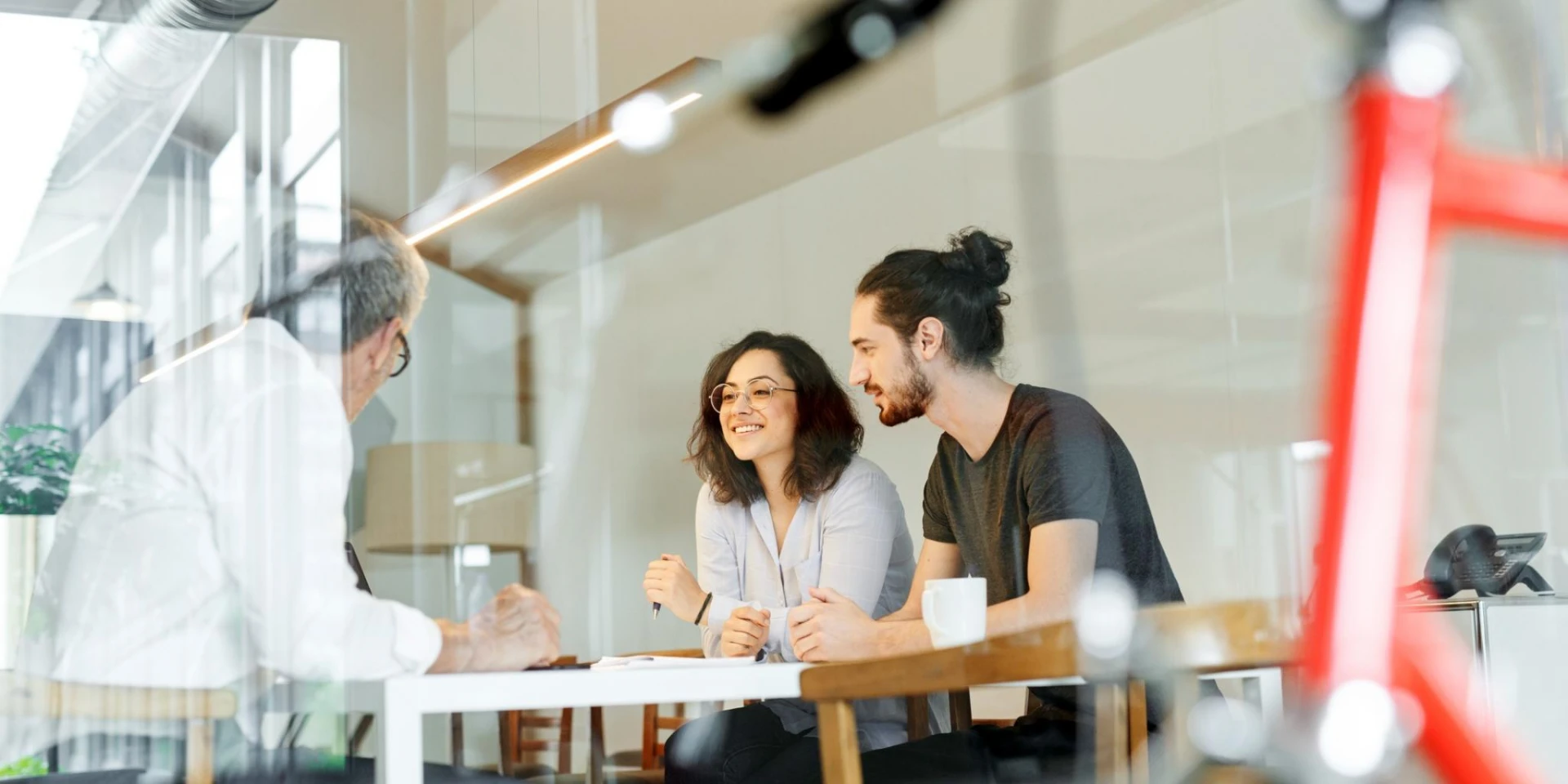 A relaxed business meeting with a man and a woman seated at a table, smiling and engaging in conversation with an older man. The setting is a modern, glass-walled office, with a red bicycle partially visible in the foreground.